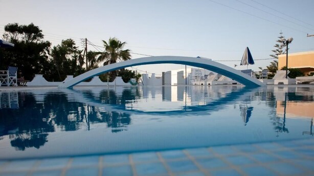 A swimming pool with a modern bridge structure, surrounded by palm trees and buildings. - THE F SUITES (KARTERADOS)