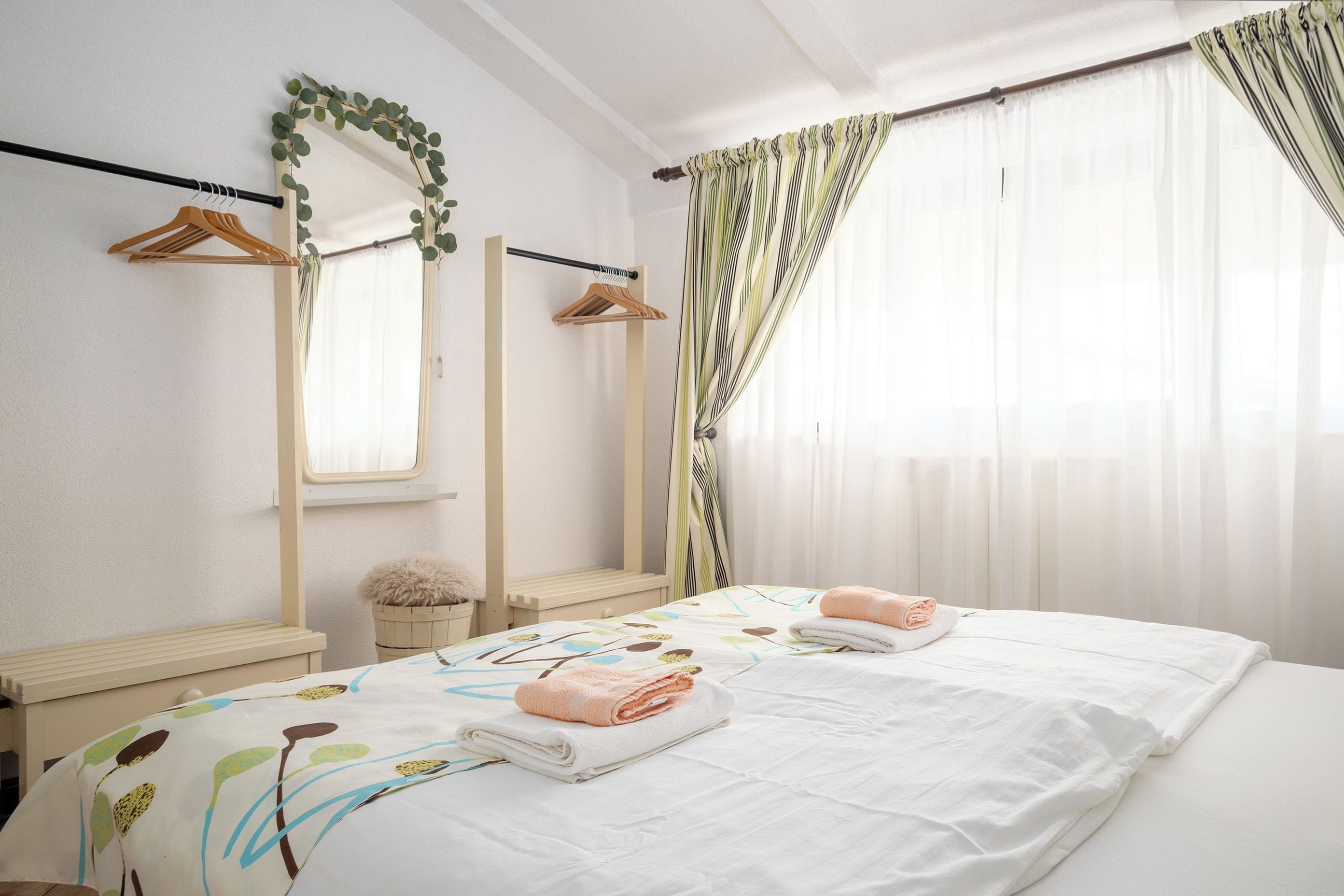 A hotel room with a bed, two bedside tables, a mirror, a hanging plant, and a window with curtains.