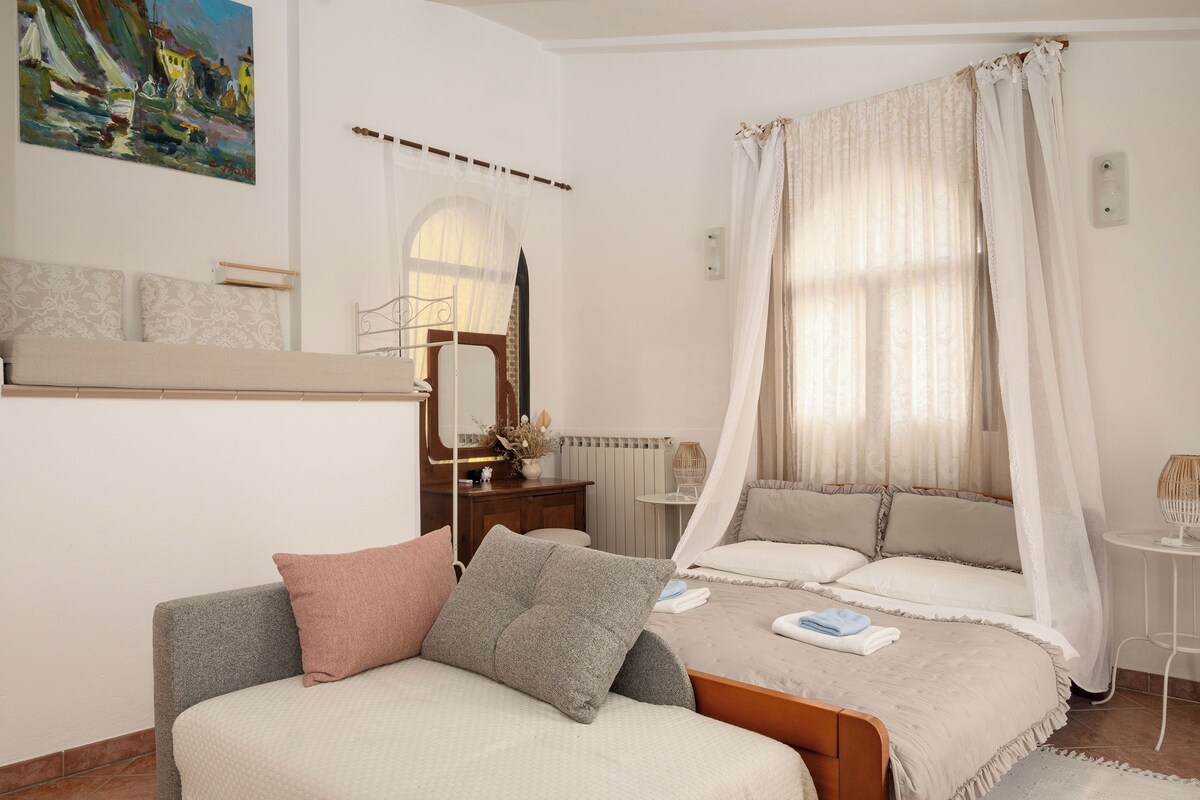Un dormitorio con cama, mesita de noche, espejo, ventana con cortinas y un cuadro en la pared.
