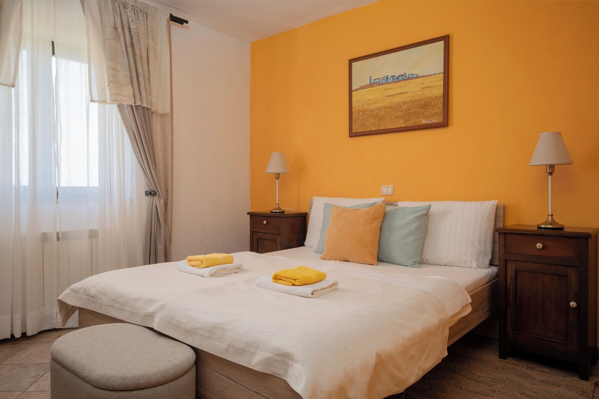 Una habitación de hotel con una cama, dos mesitas de noche, una lámpara sobre cada una, un ventanal con cortinas y un cuadro en la pared.