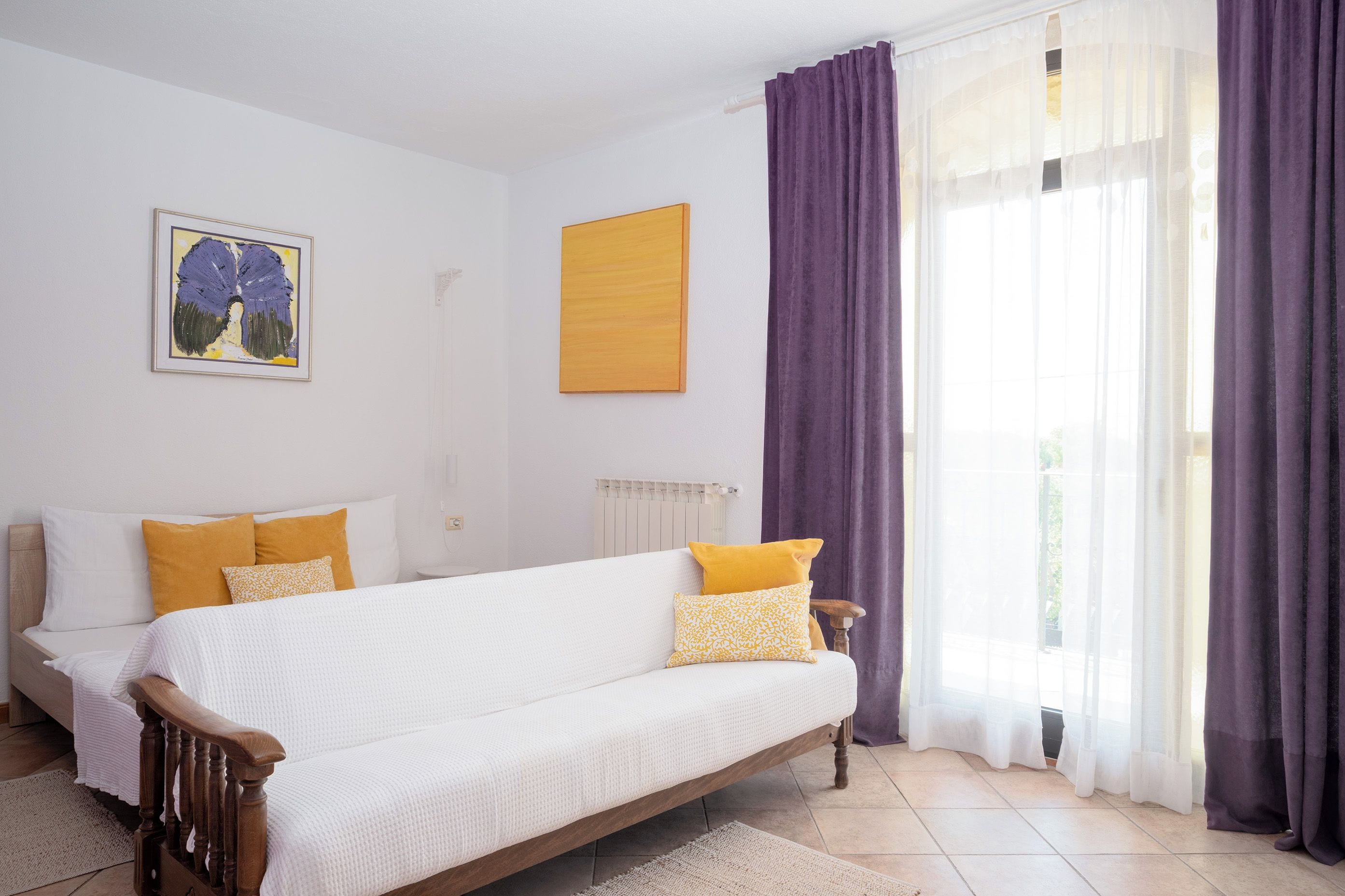 Un dormitorio con cama, una silla, un radiador, un ventanal con cortinas y un cuadro en la pared.