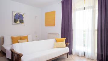 Un dormitorio con cama, una silla, un radiador, un ventanal con cortinas y un cuadro en la pared.
