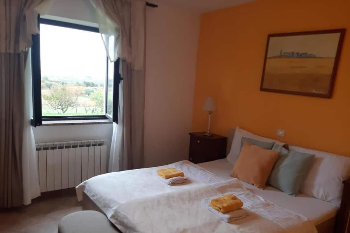 A double bed with towels, a nightstand with a lamp, a window with curtains, and a painting on the wall.