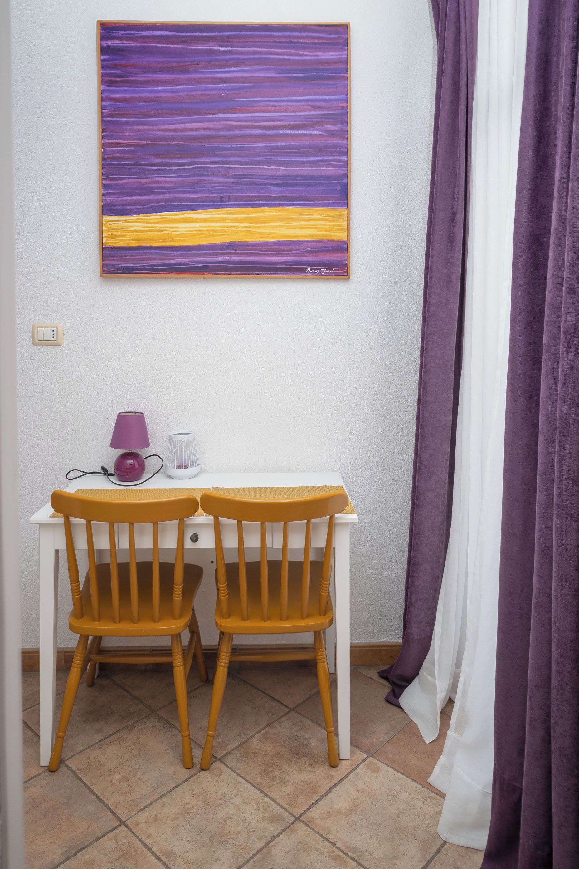 A room with a white desk, two wooden chairs, a purple lamp, and a purple curtain.