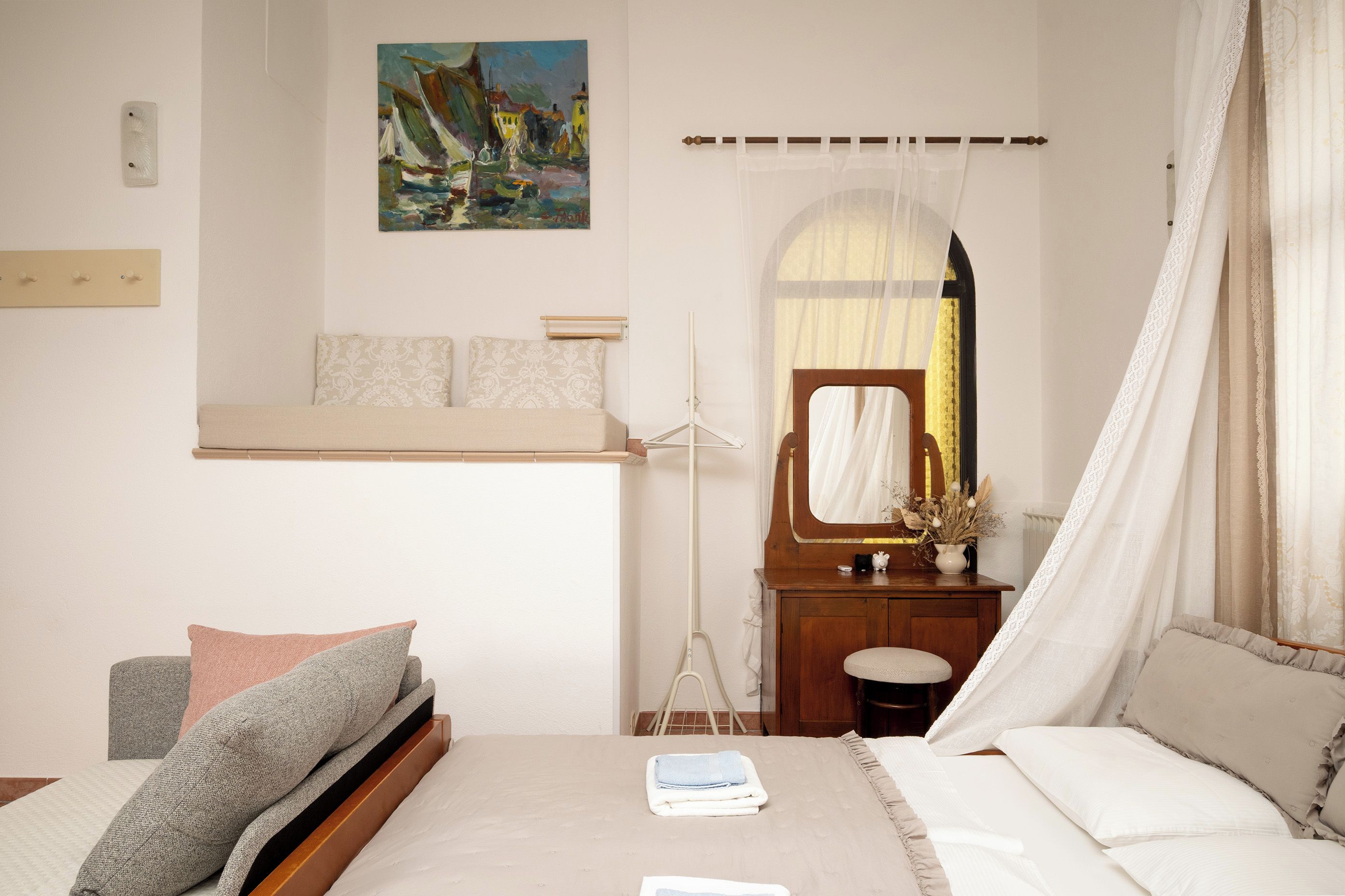 Un dormitorio con cama, mesita de noche, espejo, cuadro en la pared y ventana con cortinas.