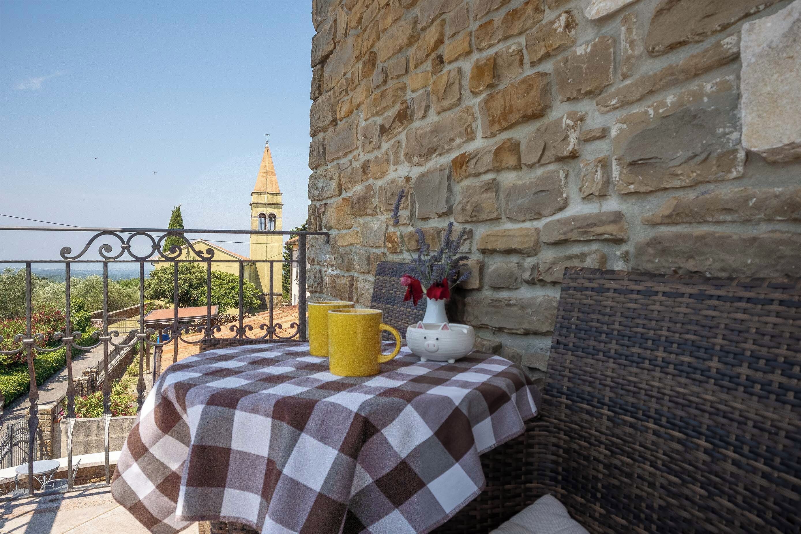 Ein Balkon mit kariertem Tischtuch, einer gelben Tasse, einem Schweinchen-Kerzenhalter und einer Vase mit Blumen.