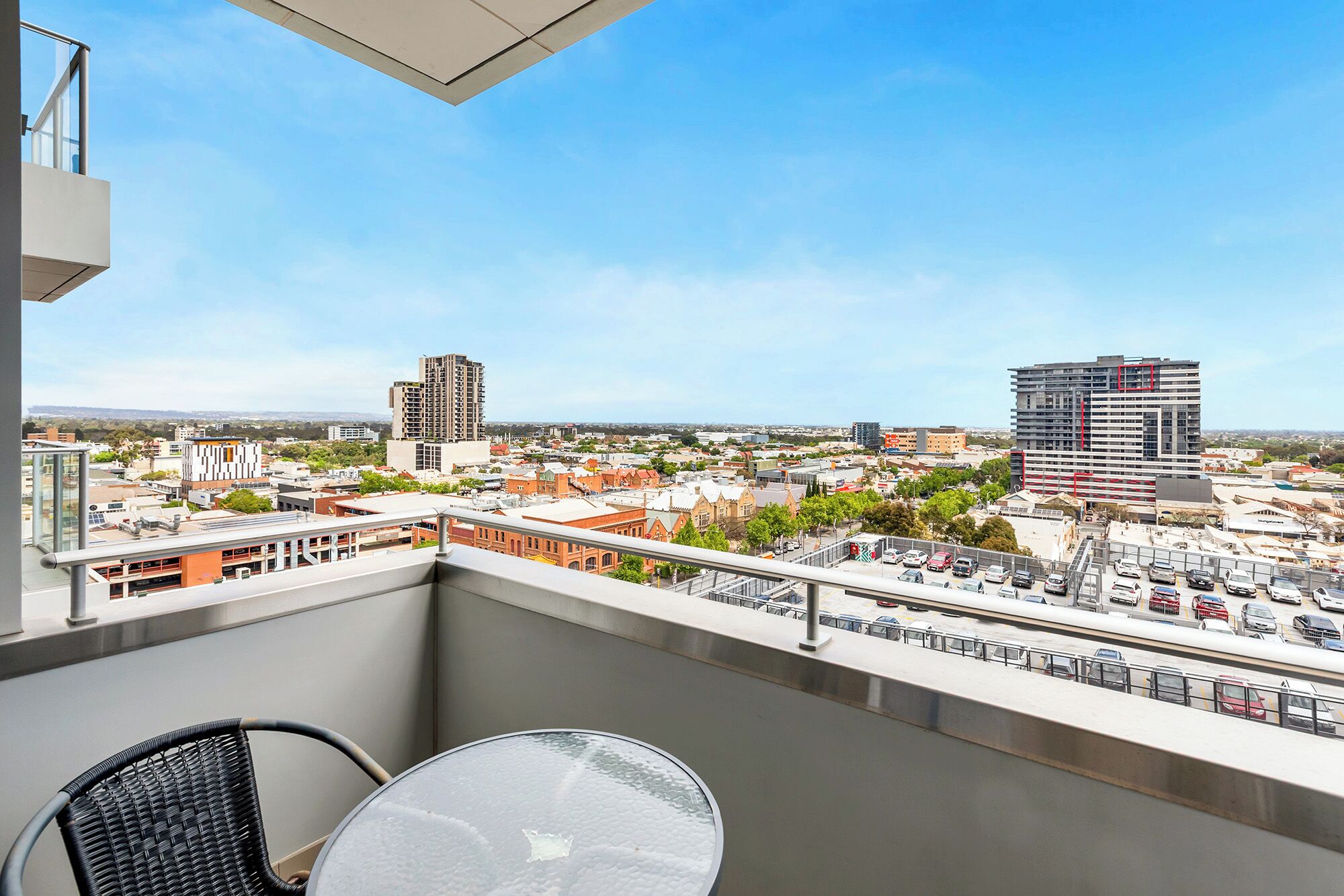 Varanda com mesa e cadeira, vista para paisagem urbana com edifícios e um estacionamento.