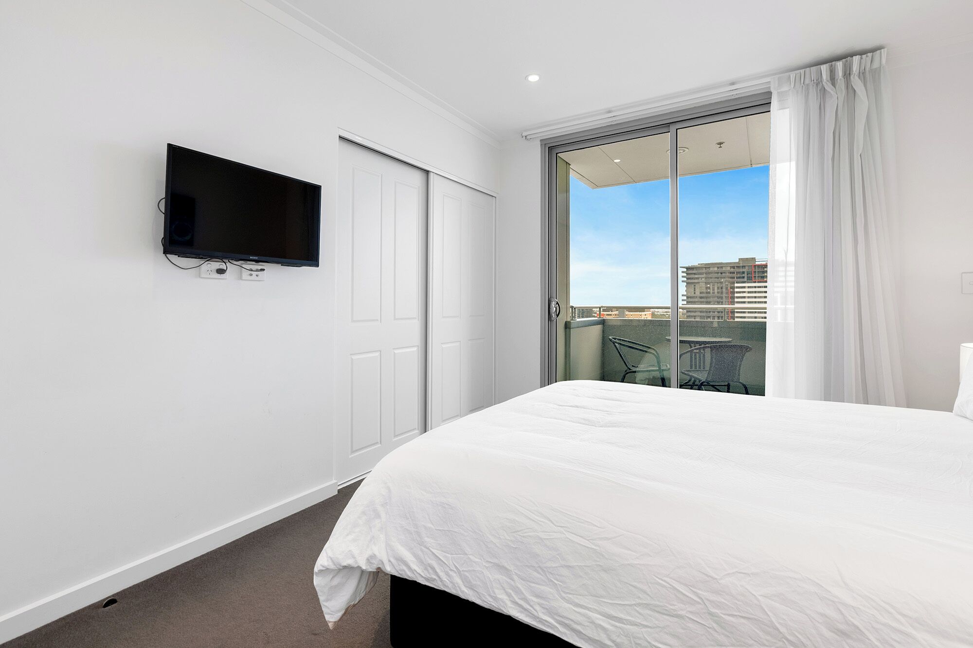 Ein Hotelzimmer mit Bett, einem an der Wand befestigten Fernseher, einer Schiebeglastür zu einem Balkon und Blick auf die Stadtlandschaft.