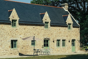 Ein Gebäude aus Stein mit einem Ziegeldach und mehreren Fenstern, vor der Fassade ein Tisch mit Stühlen.