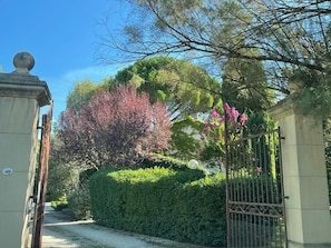 Une entrĂ©e fermĂ©e par un portail, avec un chemin menant Ă un bĂątiment, le tout entourĂ© dâune vĂ©gĂ©tation luxuriante et dâarbres en fleurs.