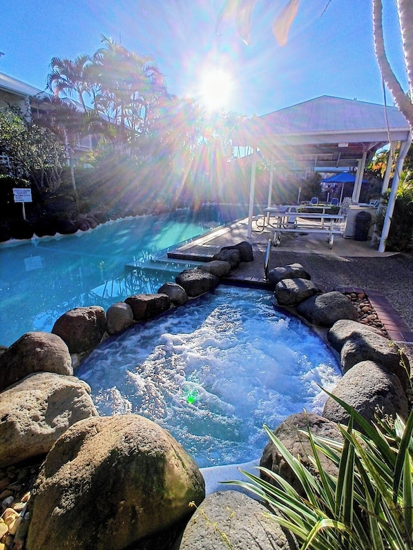 A hot tub surrounded by rocks and palm trees, with a sunlit sky and a covered seating area in the background.