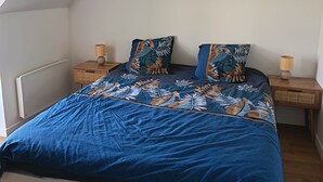 Ett sovrum med ett sluttande tak, en säng med ett blått mönstrat överkast, två matchande kuddar, en flätad golvlampa och ett sängbord med en lampa.