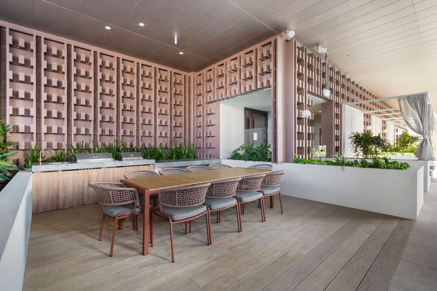 A modern outdoor dining area with a wooden table and wicker chairs, surrounded by a patterned wall and greenery.