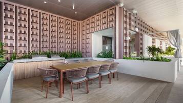 A modern outdoor dining area with a wooden table and wicker chairs, surrounded by a patterned wall and greenery.