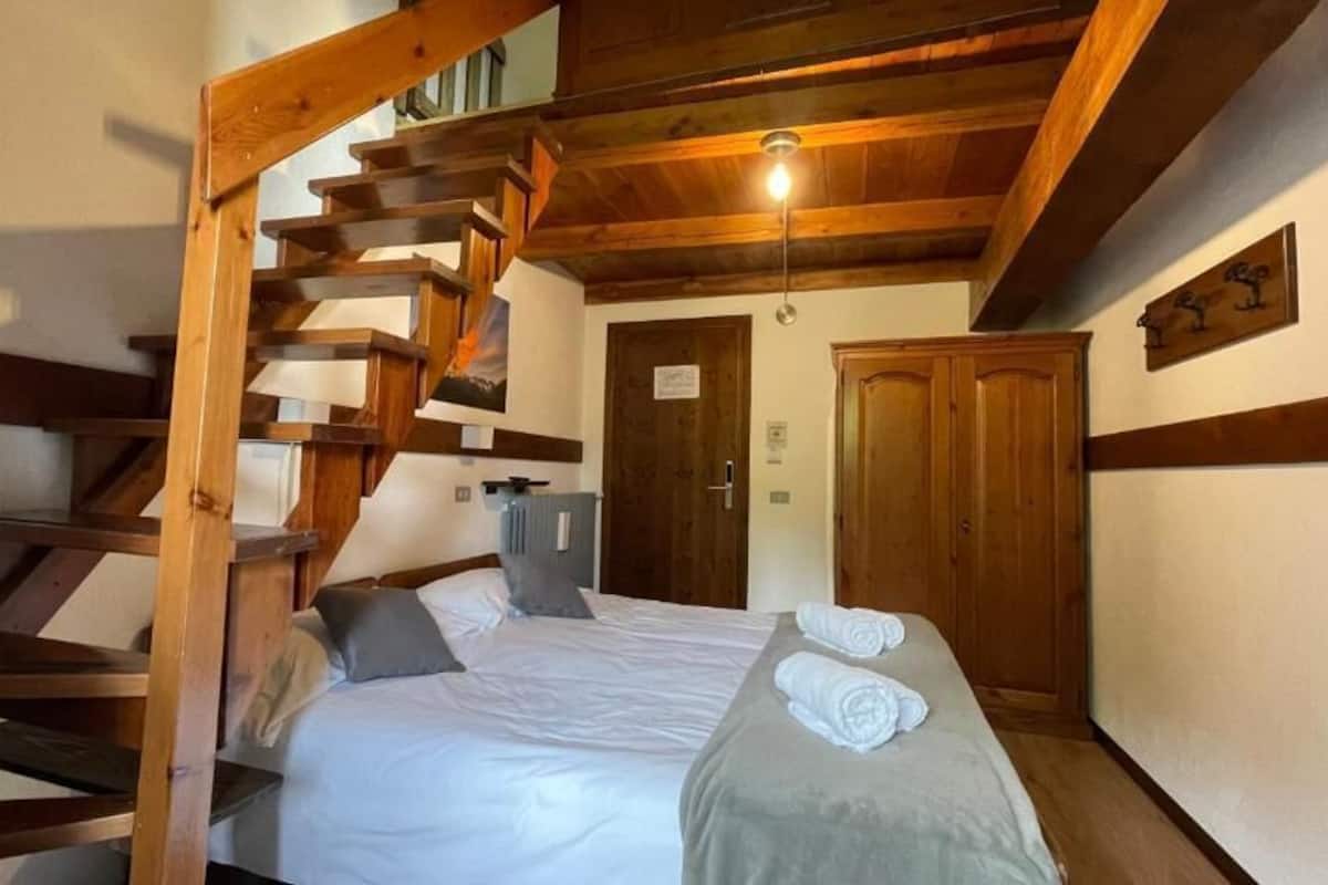 Une chambre avec un escalier en bois, un lit recouvert de draps blancs, une porte en bois et un luminaire au plafond.