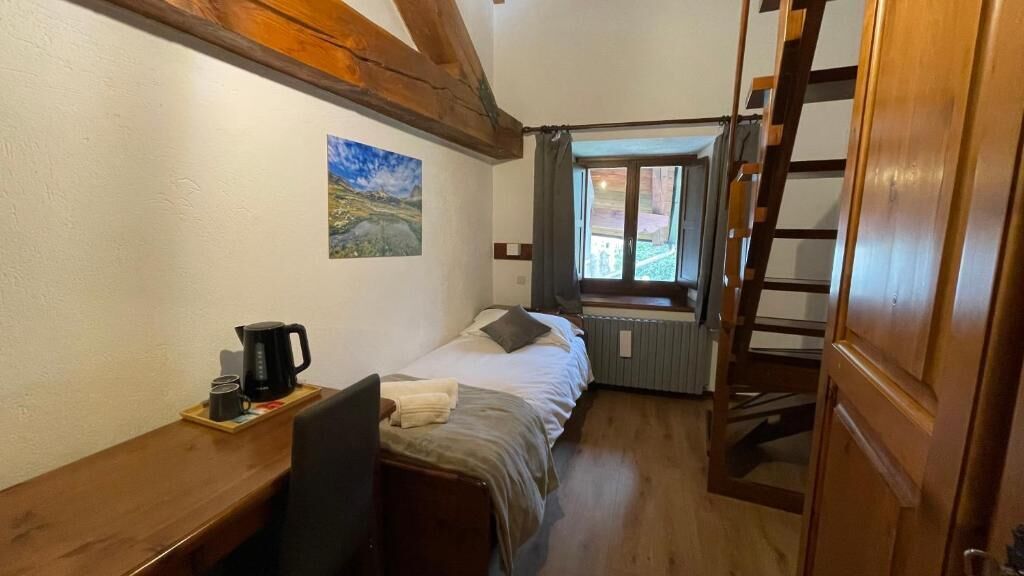 A room with a bed, a desk with a chair, a coffee maker, a window with curtains, and a painting on the wall.