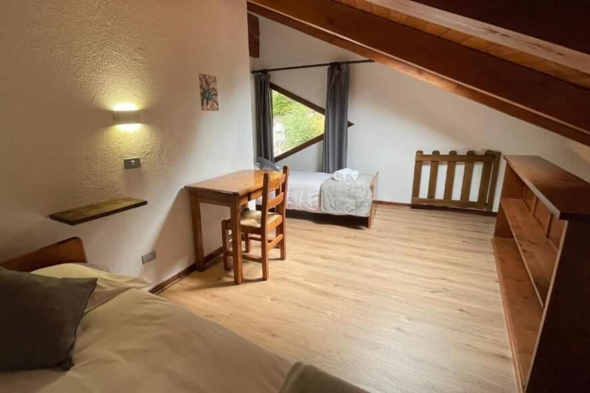 A bedroom with a bed, a desk, and a chair, a window with curtains, and a wooden ceiling.