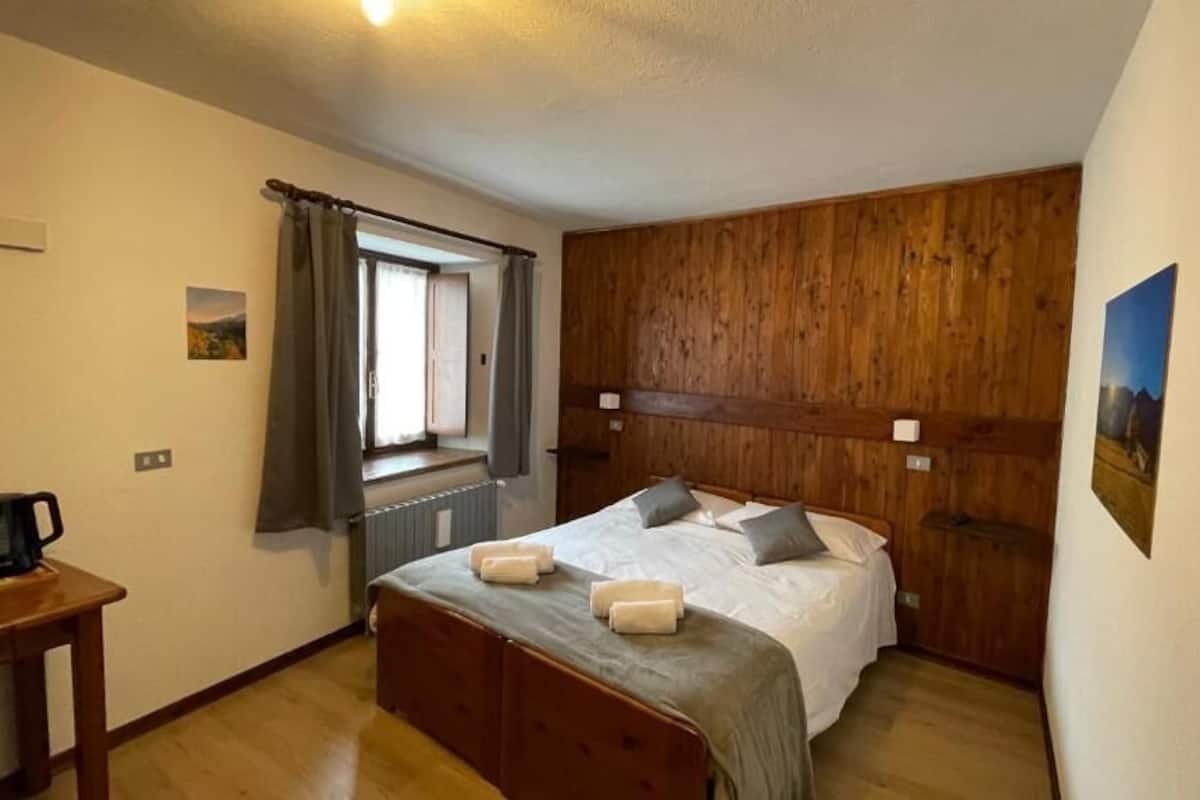 Une chambre avec une tête de lit en bois, un lit avec des oreillers, une fenêtre avec des rideaux, un radiateur et une petite table sur laquelle se trouve une cafetière.