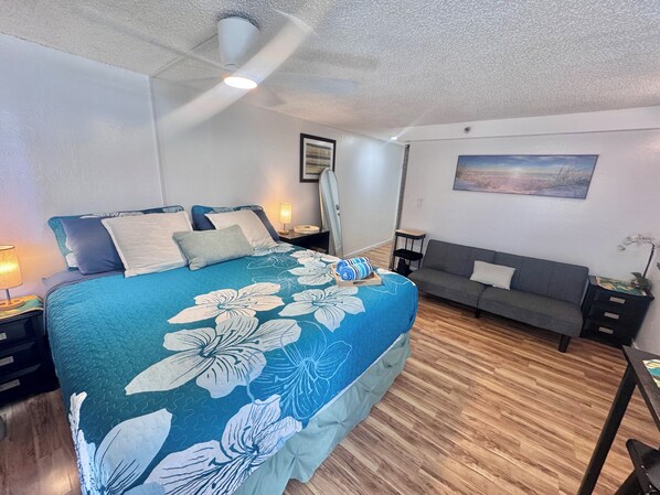 Un dormitorio con cama, mesita de noche, lámpara, sofá, un cuadro en la pared y un ventilador de techo.