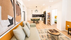 Ein modernes Wohnzimmer mit einer Couch, einem Couchtisch und einem Essbereich mit Stühlen. Im Hintergrund ist eine Küche mit Kühlschrank und Spüle zu sehen.