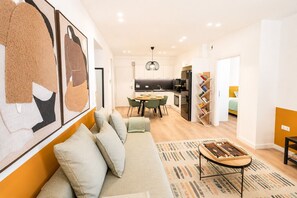 Ein modernes Wohnzimmer mit einer Couch, einem Couchtisch und einem Essbereich mit Stühlen. Im Hintergrund ist eine Küche mit Kühlschrank und Spüle zu sehen.