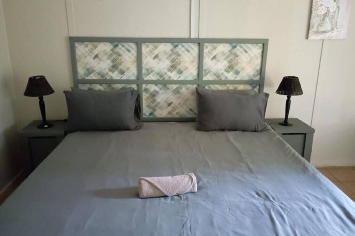 Una cama bien hecha con una colcha gris, dos almohadas y una toalla doblada. Hay dos mesitas de noche con lámparas a cada lado de la cama.