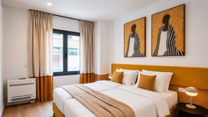 Una camera d'albergo con un letto, un comodino, una lampada, una finestra con tende e opere d'arte alle pareti.