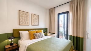 A hotel room with a bed, a nightstand, a lamp, a window with curtains, and two framed pictures on the wall. - LIV - Upscale Athenian Apartment 1C (Athens)