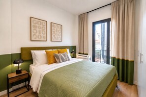 A hotel room with a bed, a nightstand, a lamp, a window with curtains, and two framed pictures on the wall. - LIV - Upscale Athenian Apartment 1C (Athens)