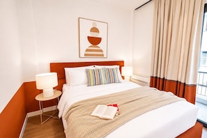 Ein Hotelzimmer mit einem Bett, zwei Nachttischlampen, einem gestreiften Kissen, einem gerahmten Kunstwerk über dem Bett und einem Fenster mit Vorhängen.