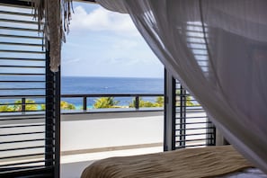 A balcony with a view of the ocean, a bed with a white bedspread, and a curtain partially drawn. - The Shore - Blue Bay Golf & Beach Resort (Sint Michiel)
