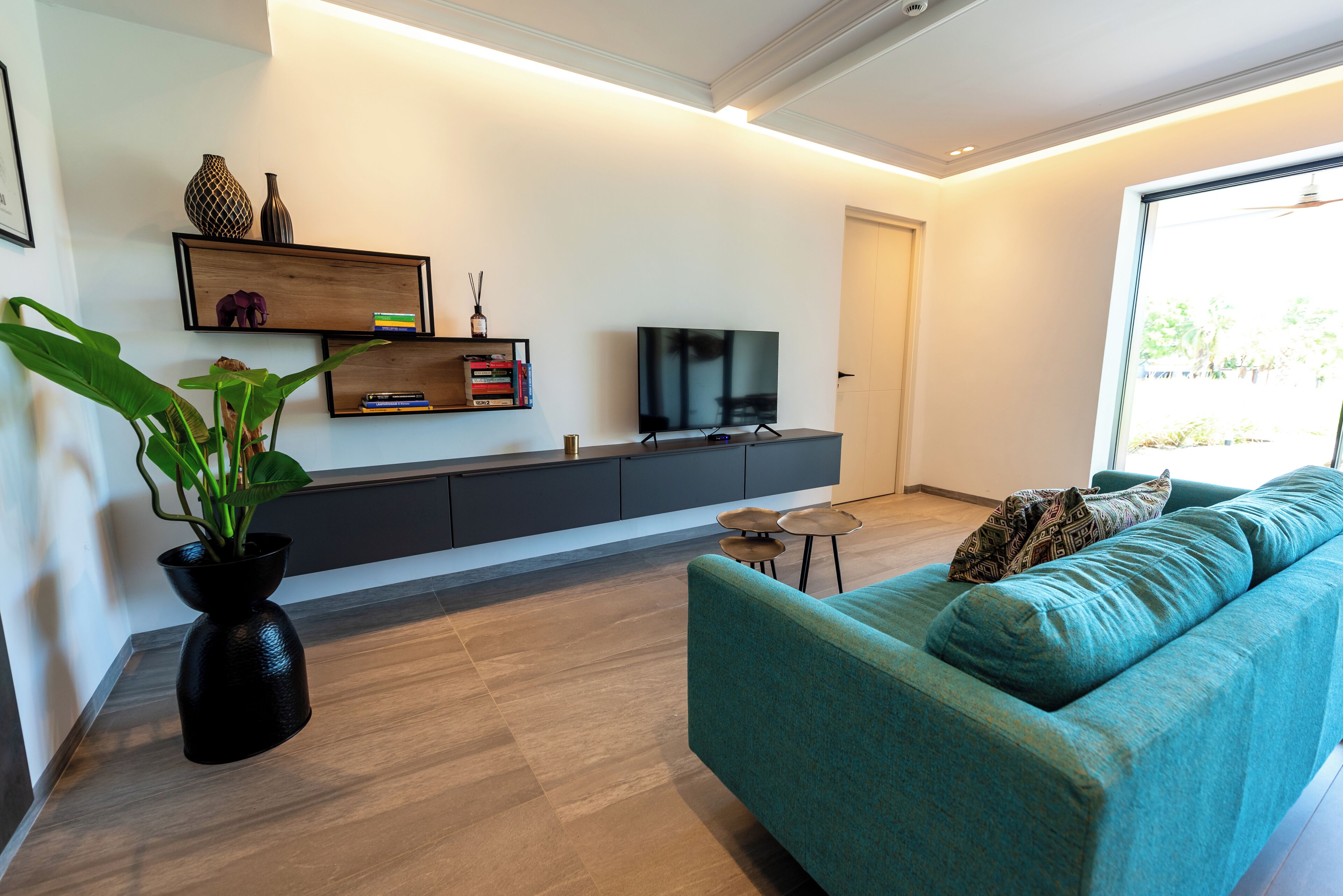 Ein modernes Wohnzimmer mit einem salbeigreenen Sofa, einem Flachbildfernseher, einer Pflanze und einem Bücherregal.