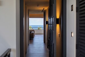A corridor leading to a balcony with a view of the sea, a sofa, and a door with a light fixture.