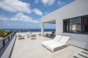 Un área de descanso al aire libre, moderna, con mobiliario blanco, vista al mar y cielo despejado.