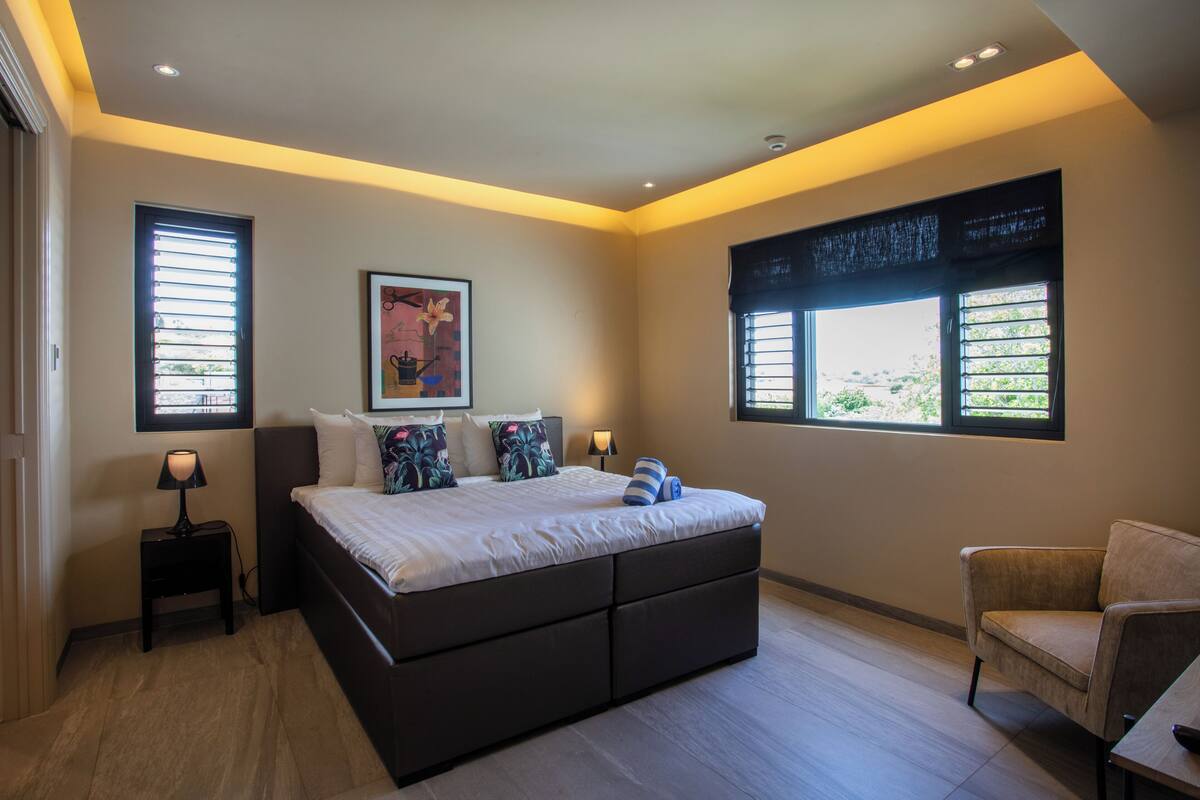 Un dormitorio con cama, mesitas de noche, una silla y ventanas amplias.