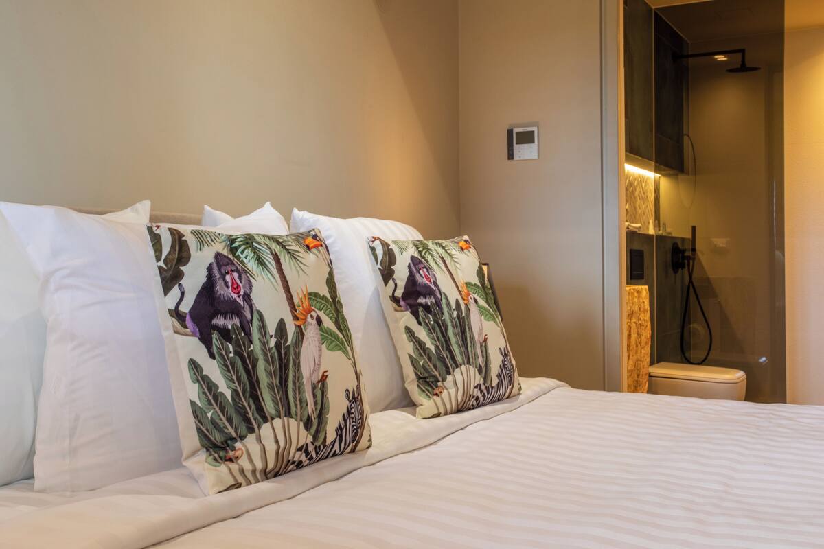 Habitación de hotel con cama, almohadas con estampados tropicales, zona de ducha y panel de control en la pared.