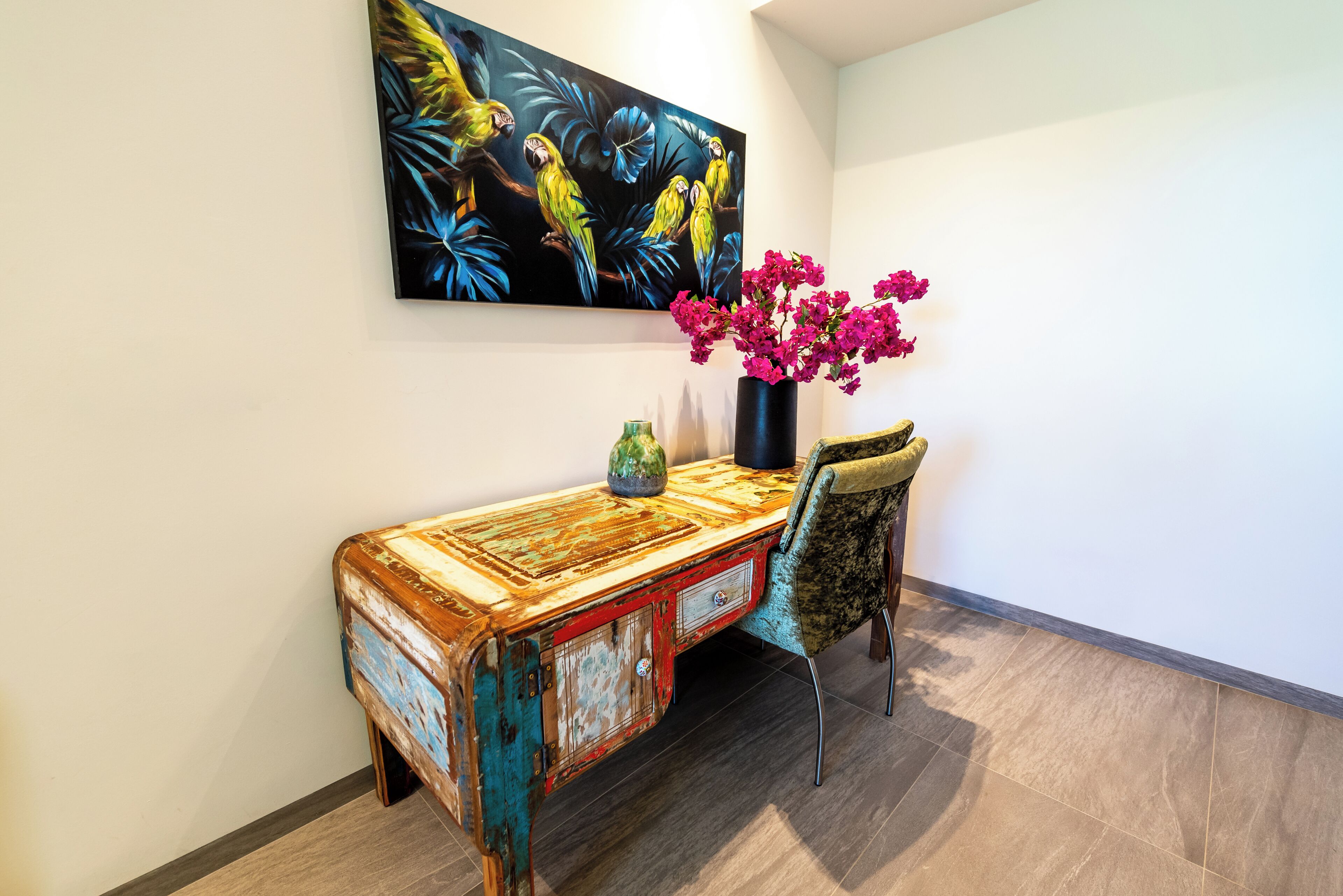A vintage-style desk with a vase of pink flowers, a chair, and a painting of parrots on the wall.