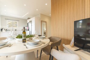 A modern dining area with a round table set for a meal, a television on a stand, and a kitchen in the background.