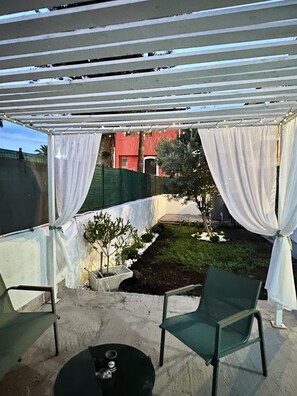 Zona exterior coberta amb una pèrgola, un jardinet i una zona de seients amb cadires i una taula.