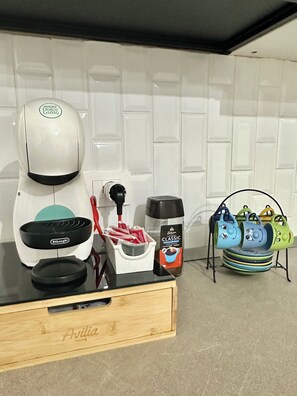 Una cuina amb cafetera, molinillo de cafè, pot de cafè i una pila de tasses de colors sobre un suport.