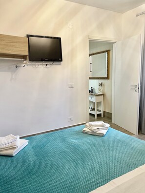 Un dormitori amb un llit, una televisió penjada a la paret, un bany visible per una porta oberta i una tovallola sobre el llit.