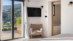 Ein Schlafzimmer mit großem Fenster, einem an der Wand montierten Fernseher, einem Sessel und einem Bett.