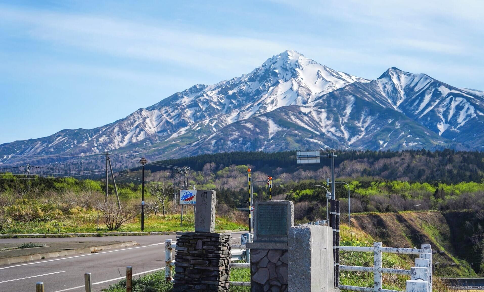A road with a stone structure and a metal fence in the foreground, a mountain with snow in the background, and a clear sky.