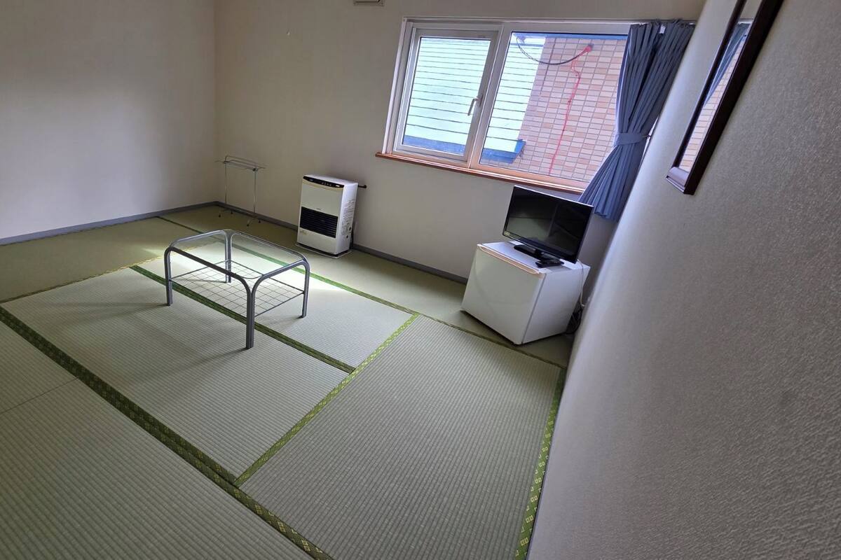 Quarto com piso de tatame, cama futon, aquecedor, suporte para TV e janela com persianas.