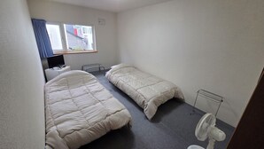 Une petite pièce, peu meublée, comprenant un lit, une table de chevet, une chaise, un bureau, un ordinateur, une fenêtre avec des rideaux et un ventilateur sur pied.