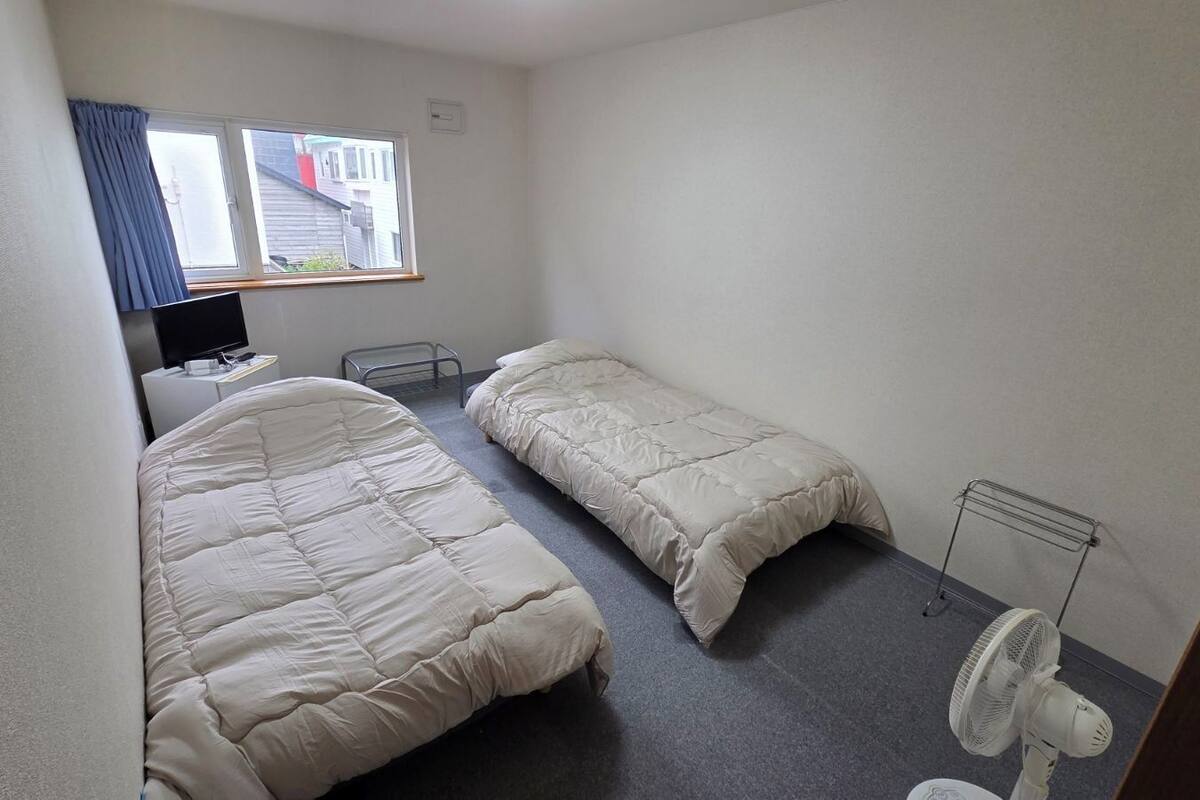 Quarto pequeno e com poucos móveis, contendo cama, uma mesa de cabeceira, cadeira, escrivaninha, computador, janela com cortinas e um ventilador.