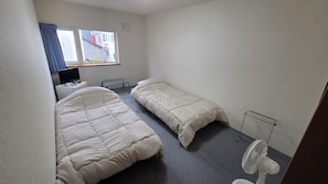 Una habitación pequeña y poco amueblada con cama, una mesita de noche, una silla, un escritorio, un ordenador, una ventana con cortinas y un ventilador de pie.