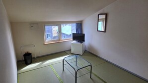 Une petite pièce avec un sol recouvert de tatamis, un téléviseur posé sur un meuble, un miroir au mur et une fenêtre avec des rideaux.