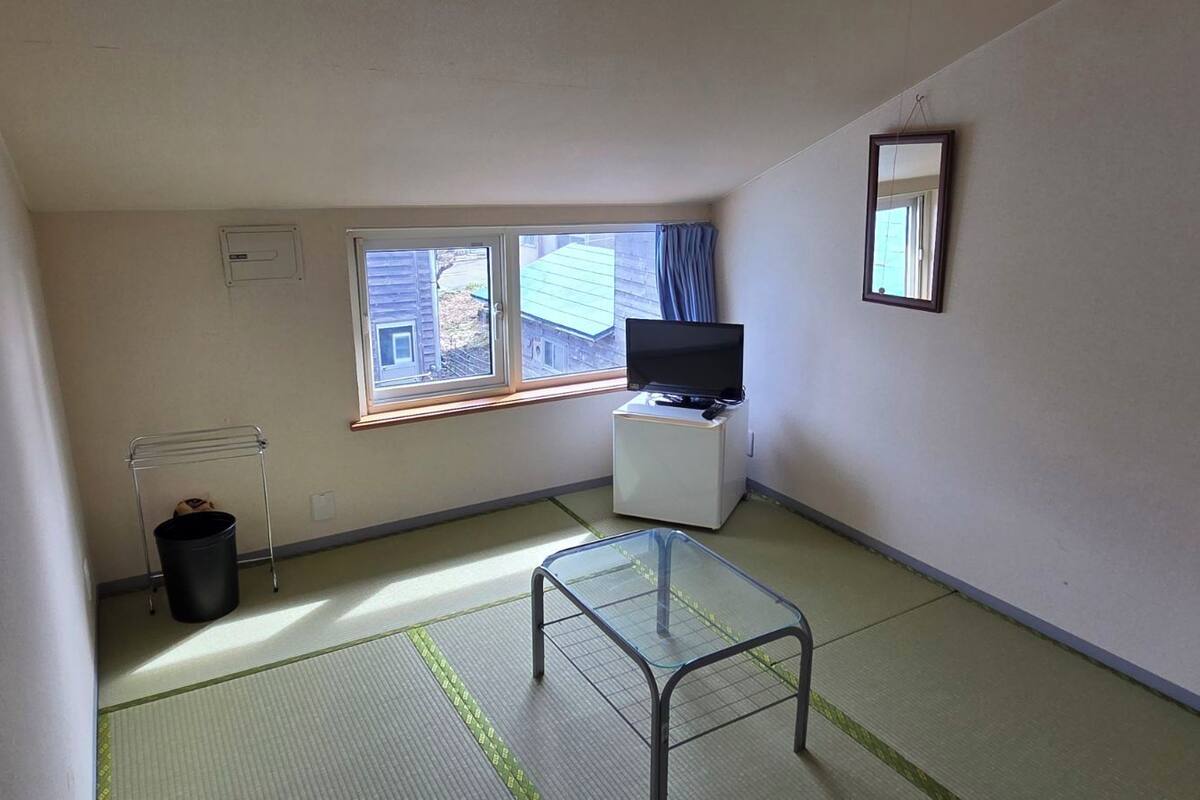 Um quarto pequeno com piso de tatame, uma televisão sobre suporte, um espelho na parede e uma janela com cortinas.