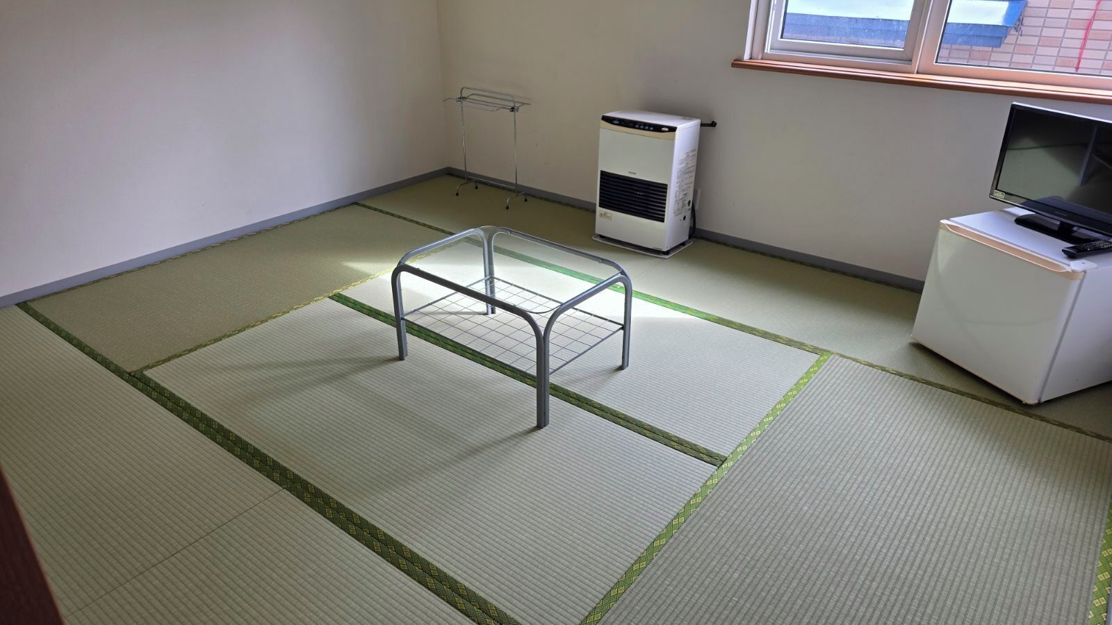 Una habitación con suelo de esteras de tatami, un taburete de metal, un calefactor, un televisor y un mueble pequeño.